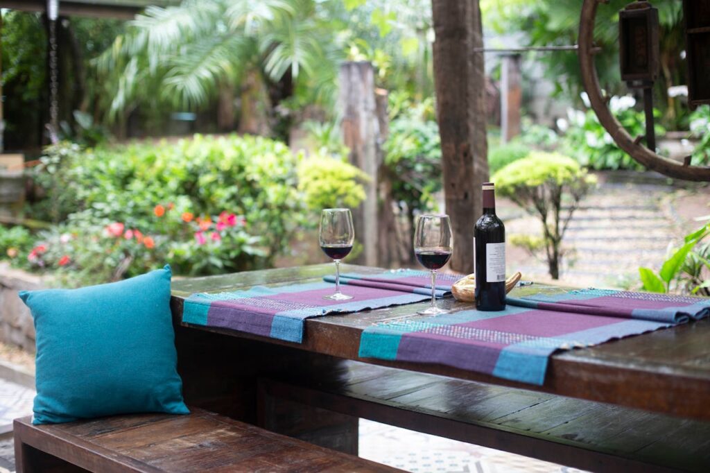 A wooden table in the outdoors with two glasses of wine and a wine bottle.